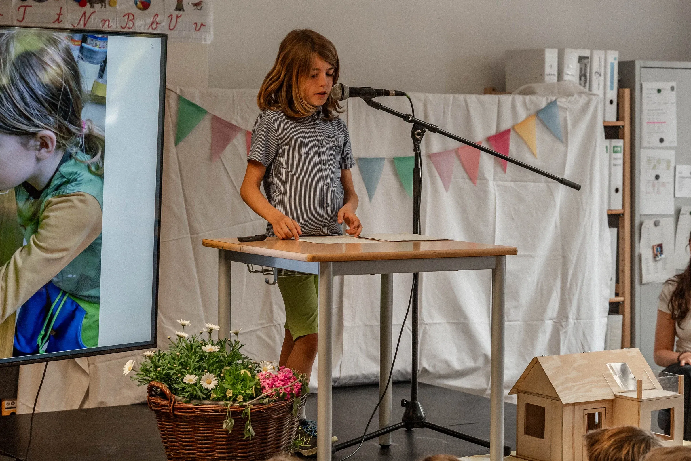 Ein Kind präsentiert seine Große Arbeit; daneben sind ein Prozessfoto und das selbst gebaute Spielhaus zu sehen
