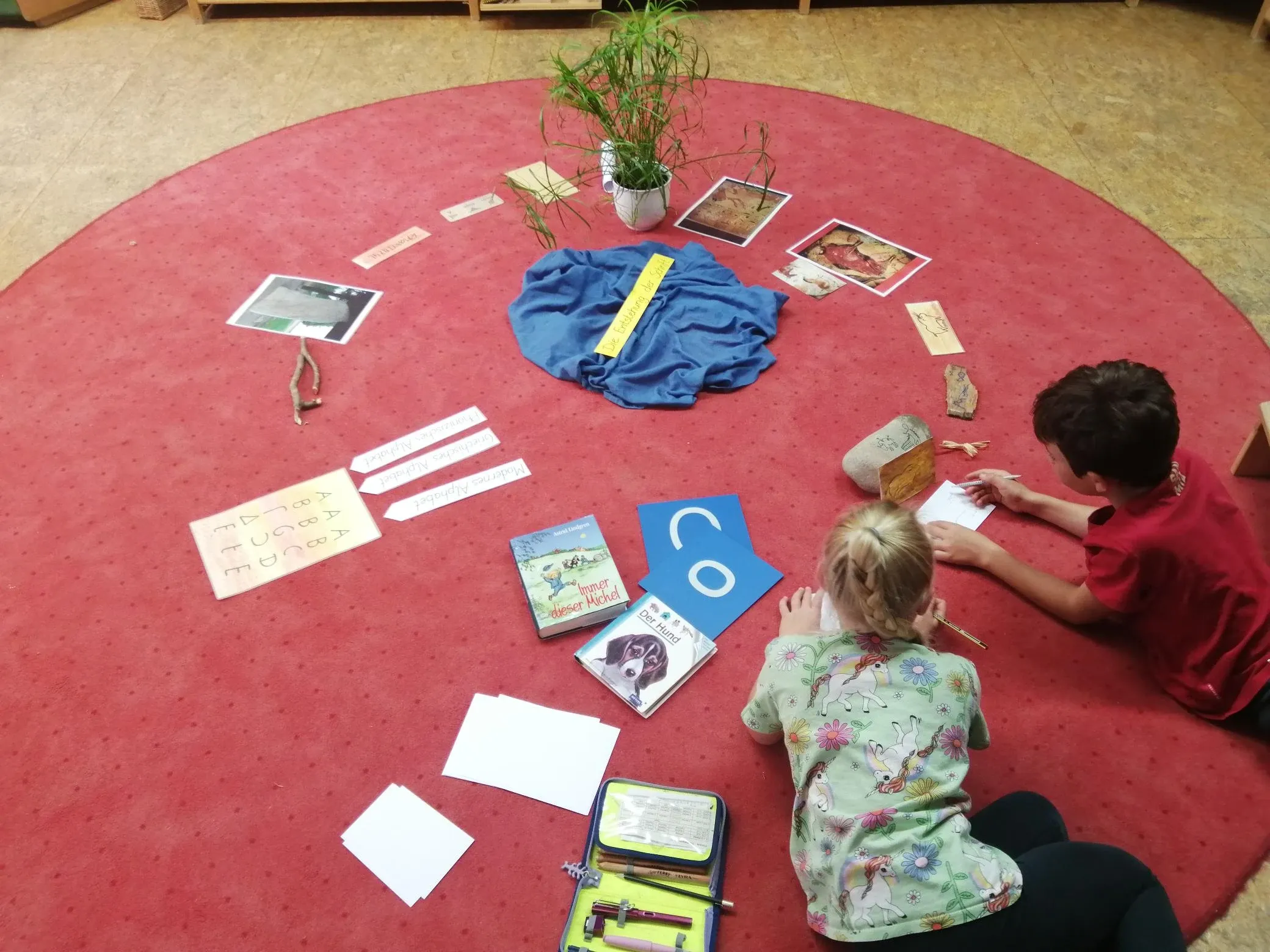 Zwei Kinder arbeiten selbstständig auf einem vorbereiteten Teppich mit Schriftzeichen, Bildkarten, Büchern und Pflanzen